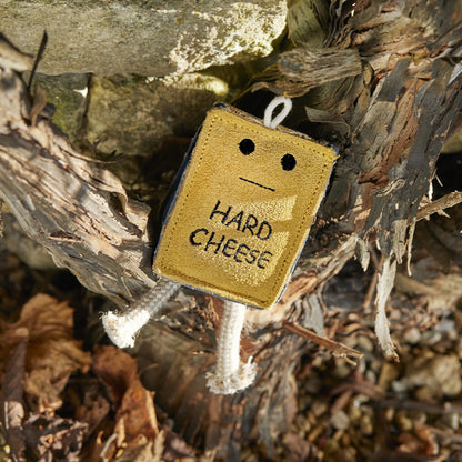 Small decorative sign shaped like a block of cheese with a face, hanging among dry leaves and branches.
