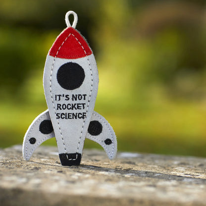 Fabric rocket-shaped ornament with 'It's Not Rocket Science' text on a blurred natural background